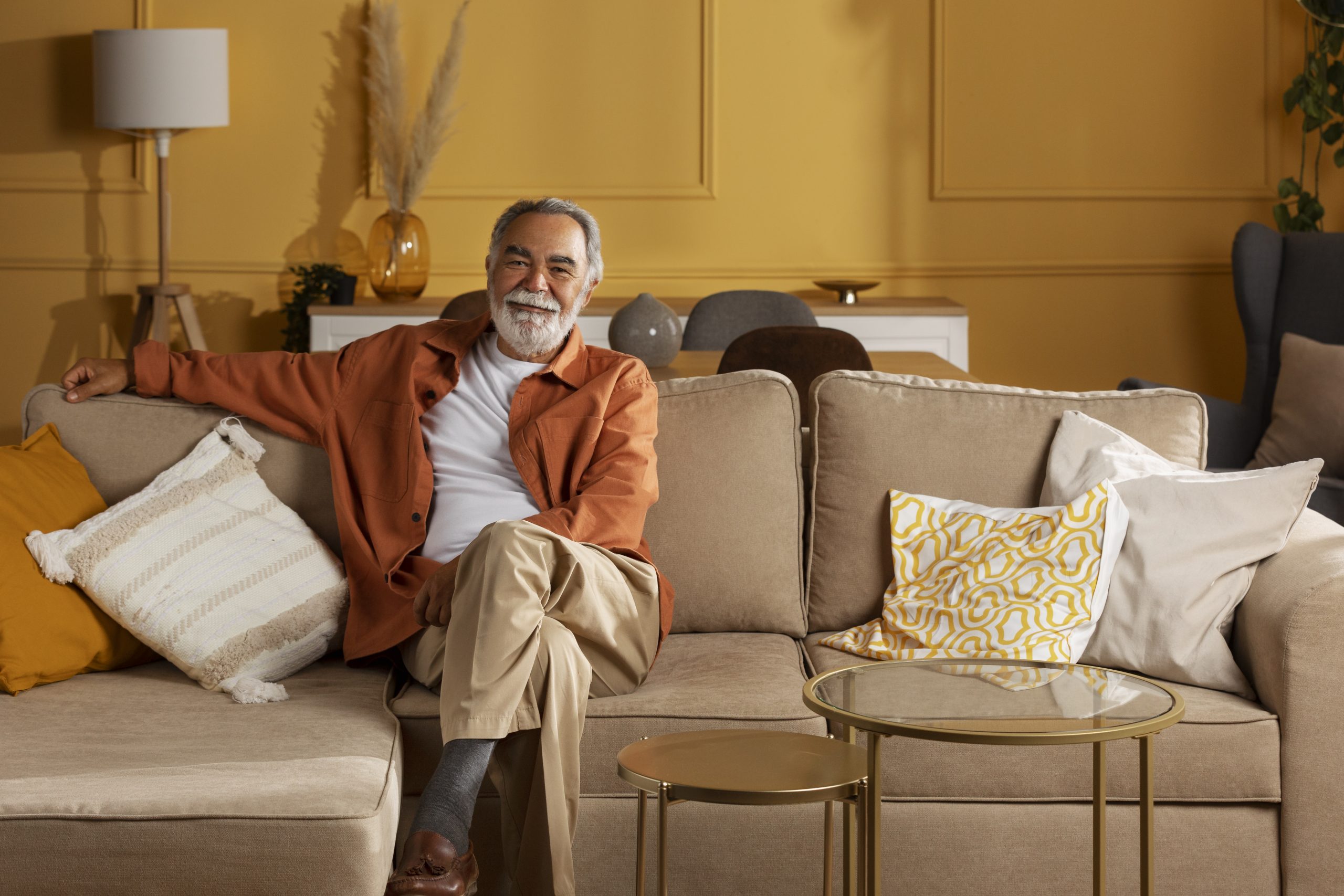 Um homem idoso sentado em um sofá bege, com as pernas cruzadas e o braço apoiado no encosto do sofá. Ele está vestido com uma jaqueta laranja, camisa branca e calça bege clara. O fundo da sala de estar tem um ambiente quente e aconchegante, com paredes amarelas suaves e decoração combinando.